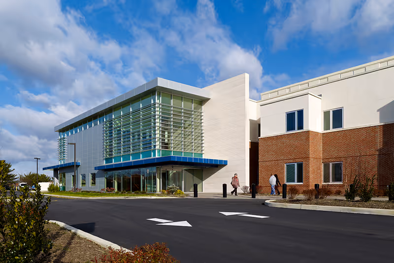 Exterior view of a modern building with a combination of glass, metal, and brick facade under a partly cloudy sky. Two people are walking near the entrance on a paved driveway with directional arrows.