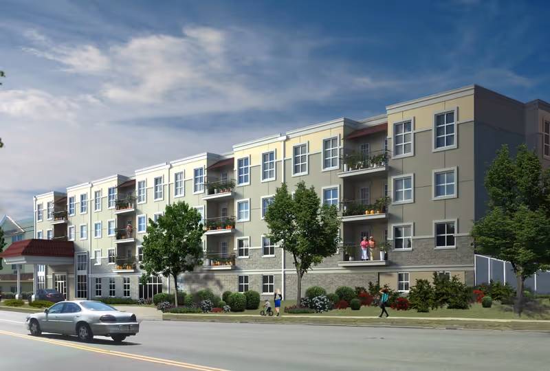 Exterior view of a multi-story residential building with balconies, trees, and landscaping along the sidewalk. Several people are visible walking or standing near the building. A car is driving on the road in front of the building under a partly cloudy sky.