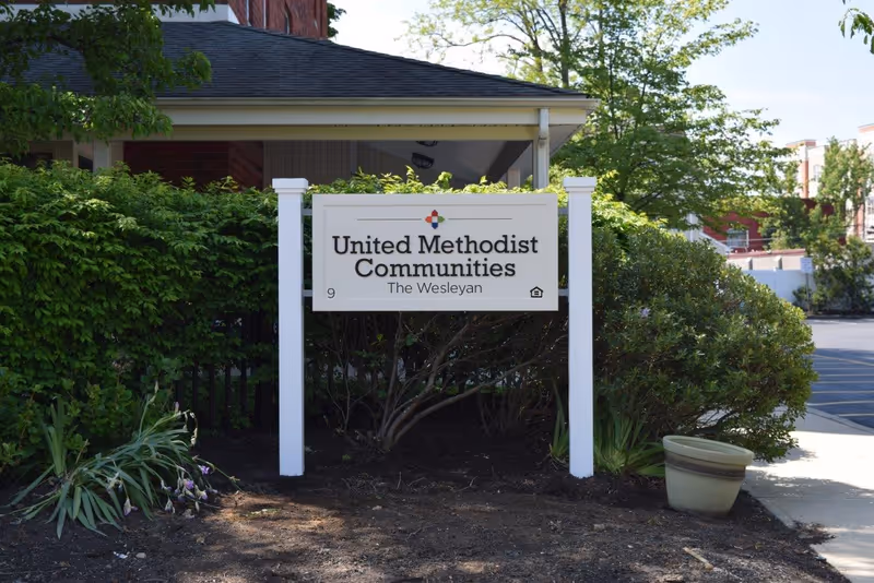 White freestanding sign reading "United Methodist Communities The Wesleyan" set among shrubs in front of a building.