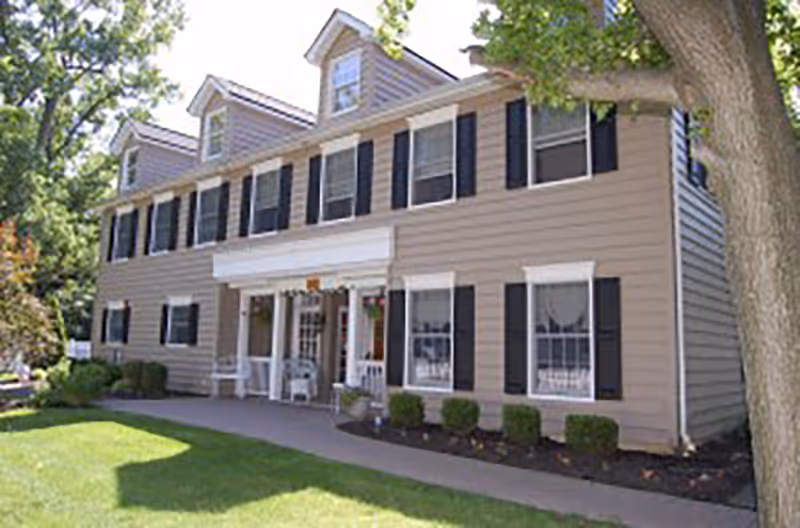 Exterior view of a two-story beige building with black shutters and white trim, featuring a covered entrance with white columns and chairs on the porch. The building is surrounded by green grass, bushes, and trees.