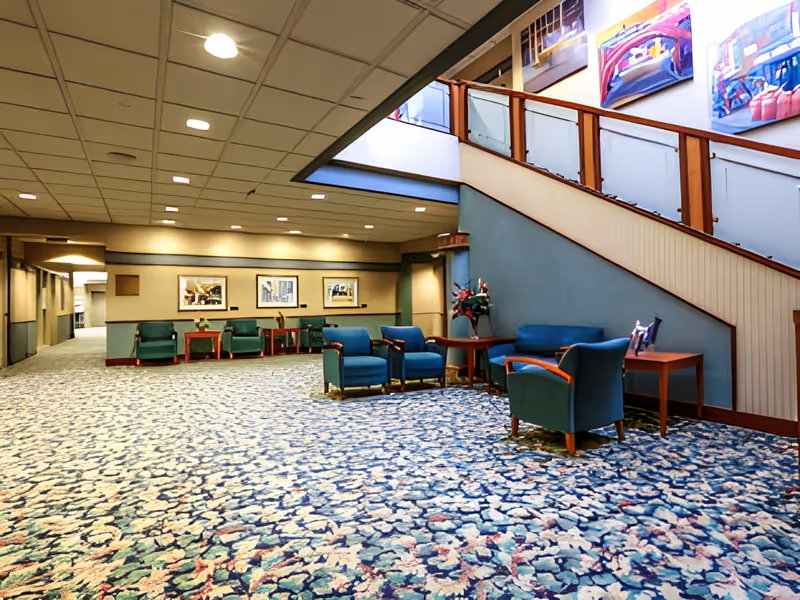 A spacious interior common area with patterned carpet, blue upholstered chairs and sofas arranged around wooden tables, a staircase with wooden railing and frosted glass panels, and framed artwork on the walls.