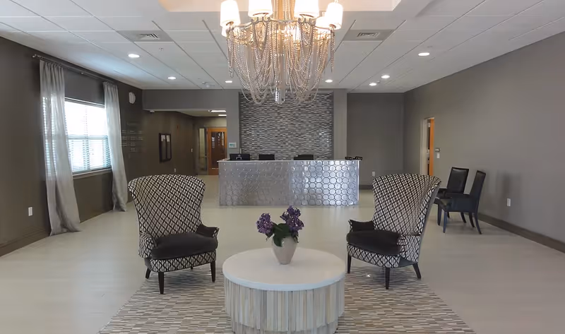 A modern reception area with two patterned armchairs facing a round white table with a vase of purple flowers. Behind the table is a reception desk with a textured front and a decorative wall. A large chandelier hangs from the ceiling, and there are windows with sheer curtains on the left side. Additional chairs are placed against the right wall.