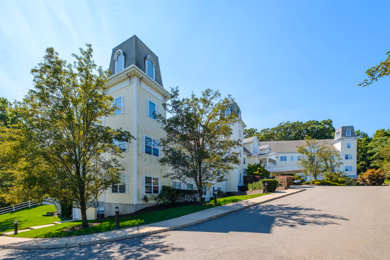 Front exterior of a multi-story senior living building with pale siding, mansard-roof towers, trees, and a driveway under a blue sky.