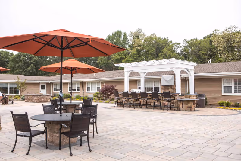 Outdoor patio area with round tables and chairs under large orange umbrellas, a white pergola with bar seating, and a building with windows and a brown roof in the background surrounded by trees.