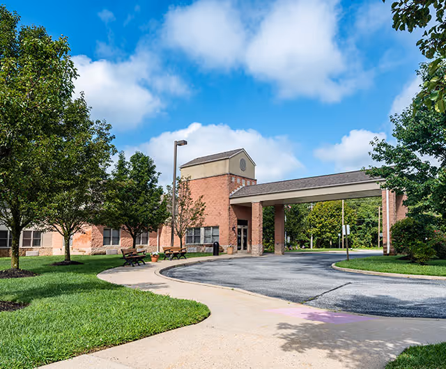 Exterior view of a senior living facility building with a covered entrance driveway. The building is made of brick with a beige upper section and surrounded by green trees and grass under a blue sky with some clouds. There are benches and a sidewalk leading to the entrance.