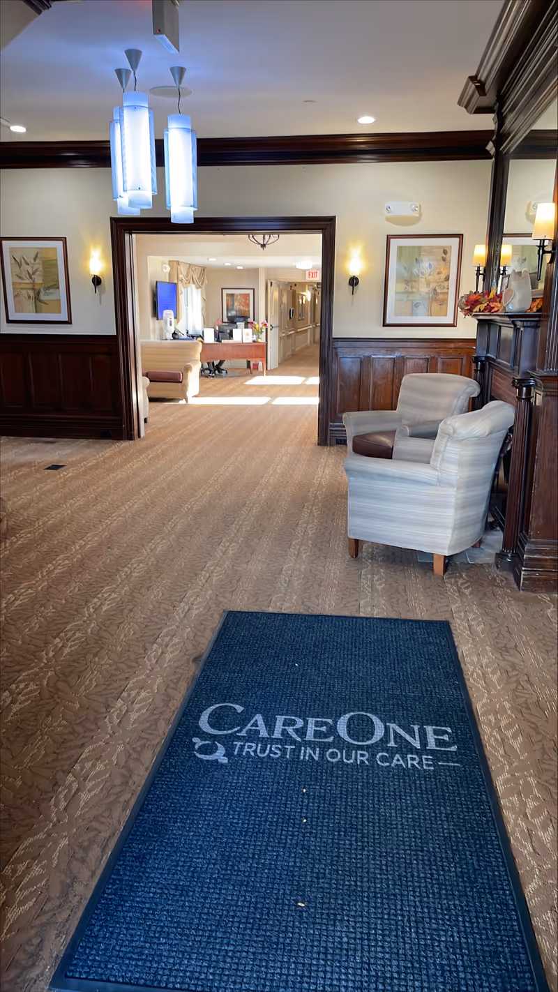 Interior view of a senior living facility hallway with carpeted floor and wooden paneling on the walls. There is a blue mat on the floor with the text 'CareOne Trust in Our Care'. Two armchairs are positioned near a wooden mantel with decorative items. The hallway leads to a reception desk area with a computer and seating. The space is well-lit with ceiling lights and wall sconces, and framed artwork hangs on the walls.