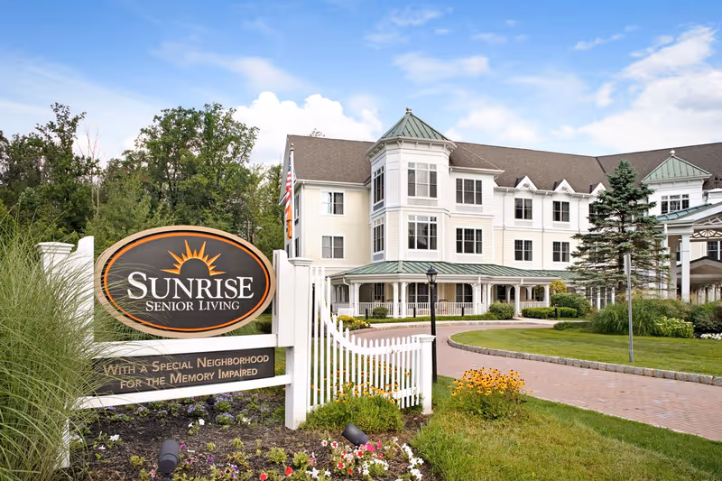 Exterior view of Sunrise Senior Living facility with a white multi-story building featuring green roofs and a wraparound porch. In the foreground, there is a landscaped garden with flowers and a sign that reads 'Sunrise Senior Living - With a Special Neighborhood for the Memory Impaired'. The sky is blue with some clouds.