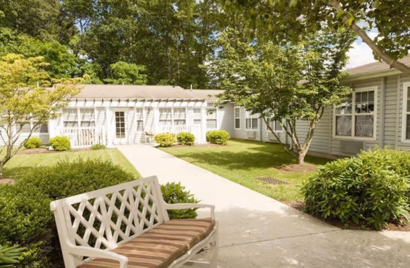 Outdoor courtyard area of a senior living facility with a white bench with a striped cushion in the foreground, surrounded by green bushes and trees. A concrete pathway leads to a light-colored building with multiple windows and a door under a pergola. The sky is partly cloudy and the area is well-maintained with green grass and landscaping.