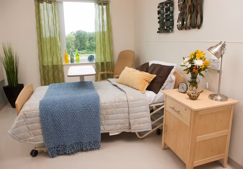 A neatly made single bed in a room with a blue textured blanket, beige and brown pillows, a wooden nightstand with a lamp, a clock, a decorative bird, and a vase of flowers. There is a window with green curtains and decorative bottles on the windowsill, a beige armchair, and a tall green plant in a black pot.