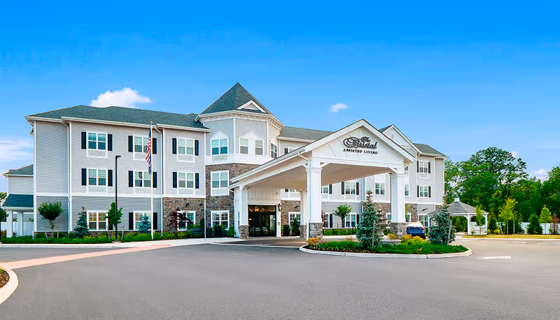 Exterior view of The Bristal Assisted Living at Somerset, a three-story building with a covered entrance, stone and siding facade, surrounded by landscaped greenery and a clear blue sky.
