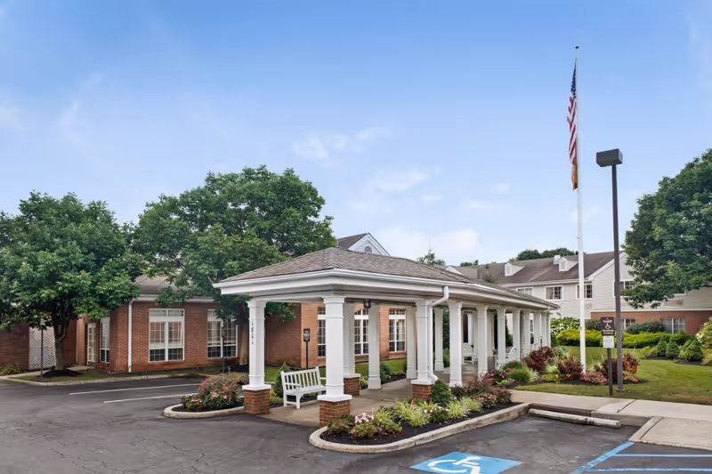 Exterior view of Brighton Gardens of Edison showing a covered entrance with white columns and benches, surrounded by landscaped greenery and trees, with an American flag on a flagpole and a clear blue sky.