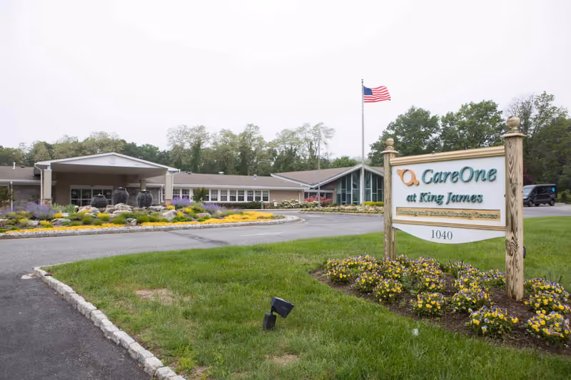 Exterior view of a senior living facility with a landscaped garden featuring colorful flowers and three large decorative urns. The building has a covered entrance and large windows. An American flag flies on a flagpole near a wooden sign that reads 'CareOne at King James Skilled Nursing and Rehabilitation Center 1040'.