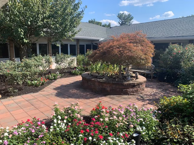 A peaceful outdoor courtyard with a circular raised brick planter containing a small tree and plants. Surrounding the planter are paved walkways and flower beds filled with colorful flowers and greenery. The courtyard is enclosed by a single-story building with windows and a gray roof under a partly cloudy blue sky.