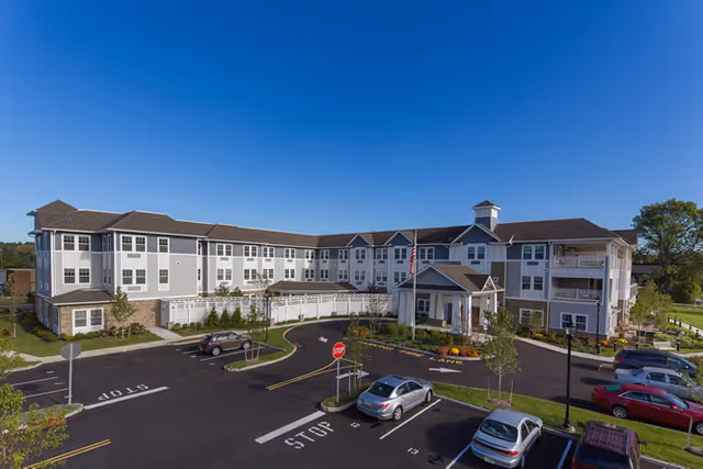 Three-story senior living building with a porte-cochere, driveway and parked cars in a landscaped parking lot under a clear blue sky.