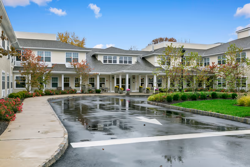 Exterior view of a senior living facility with a wet driveway, landscaped garden, and a two-story building featuring multiple windows and a covered entrance with columns. The sky is blue with a few clouds.