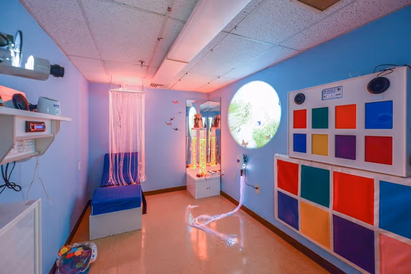 Sensory/activity room with blue walls featuring a bubble tube, fiber-optic light curtain, colorful wall panels and a round projected image.