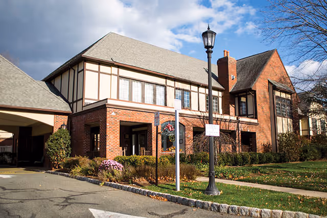 Exterior view of a two-story brick and timber senior living facility building with a covered entrance, landscaped bushes and flowers, a lamppost, and a partly cloudy sky.