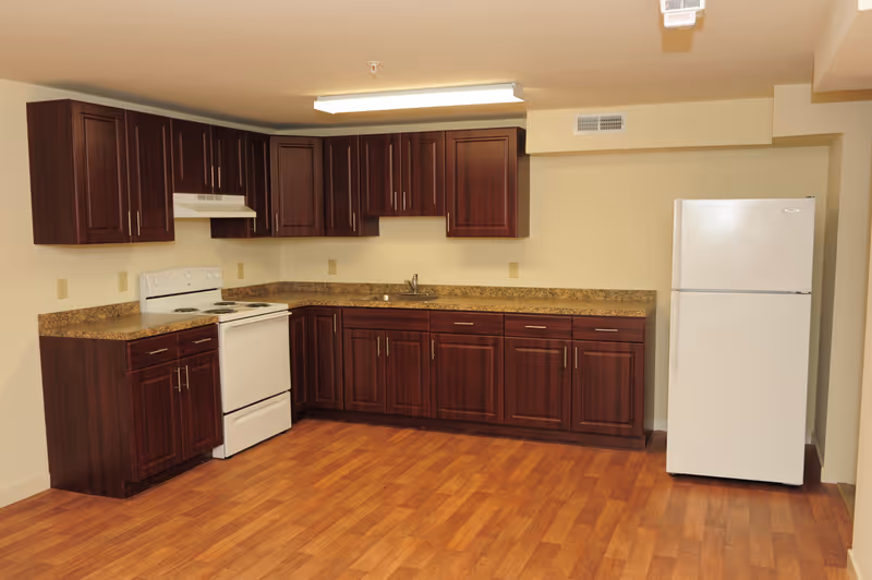 A kitchen with dark wooden cabinets, a white stove with an overhead vent, a white refrigerator, and a brown countertop. The floor is wood laminate, and the walls are painted light beige. There is a fluorescent light fixture on the ceiling.