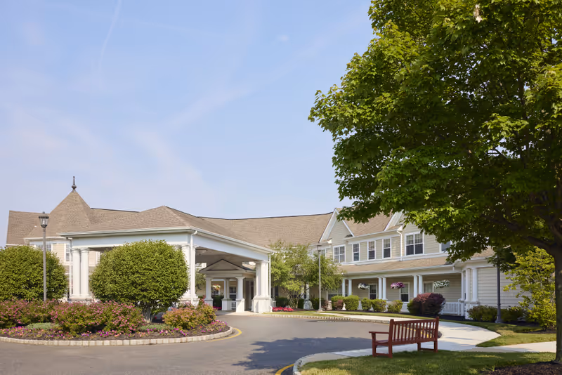 Exterior view of a senior living facility named Brandywine Wall by Monarch, featuring a large covered entrance with white columns, manicured bushes and flower beds, a tree with green leaves, a red bench on a sidewalk, and a clear blue sky.