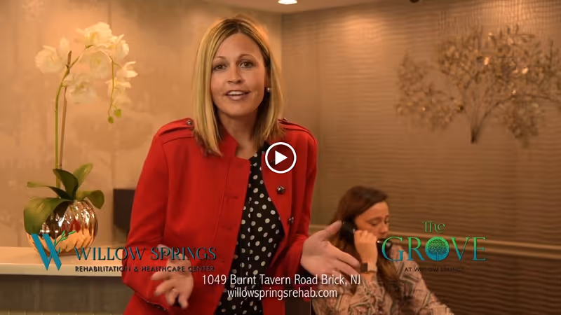 A woman in a red jacket speaking in an indoor setting with a reception desk and a potted orchid plant. Another woman is seated behind the desk, talking on the phone. The background features a textured wall with decorative elements.