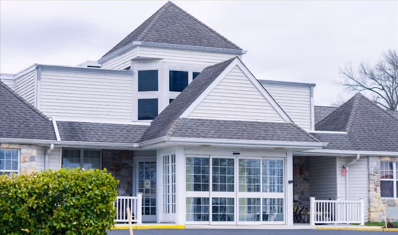 Front exterior view of a senior living facility building with a peaked roof, large windows, a glass entrance door, and a small porch area with white railings. There is a green bush in the foreground and an American flag mounted on the right side of the building.