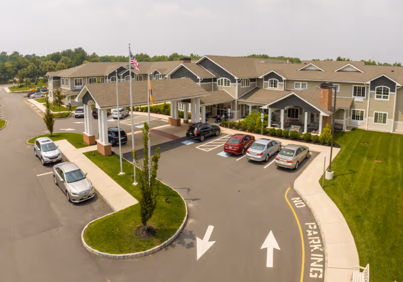 Front exterior view of Juniper Village at Hamilton, showing a large two-story building with a covered entrance, several parked cars, a flagpole with the American flag, and well-maintained landscaping including green grass and trees.