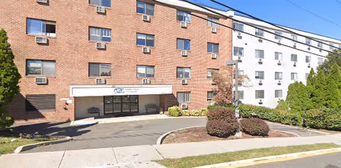Exterior view of a multi-story senior living facility with a brick and white facade, multiple windows with air conditioning units, a main entrance with a covered awning, benches near the entrance, and landscaped bushes and trees along the driveway and sidewalk.