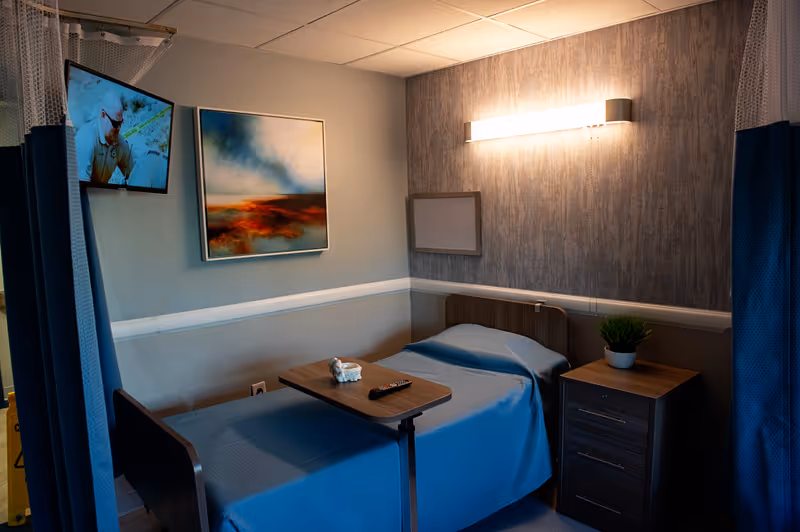 A small hospital-style room with a single bed covered in a blue blanket. A wooden bedside table with a small potted plant is next to the bed. A wall-mounted TV is on the left side, showing a man in sunglasses. Above the bed is a modern light fixture and a colorful abstract painting on the wall. A wooden overbed table holds a remote control and a small decorative item. Blue privacy curtains partially frame the room.