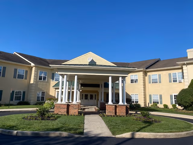 Front exterior view of a two-story senior living facility building with beige walls, white columns supporting a covered entrance, green shutters on windows, and a clear blue sky above.