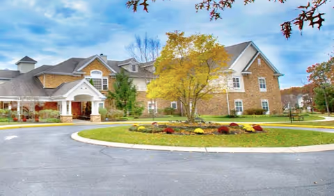 Brick multi-story senior living building with a covered porte-cochere, circular driveway and landscaped lawn with trees and flowers.