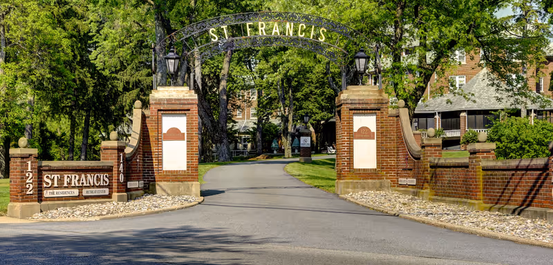 Brick entrance gate and arched sign reading 'St Francis' opening onto a tree-lined driveway toward residential buildings.