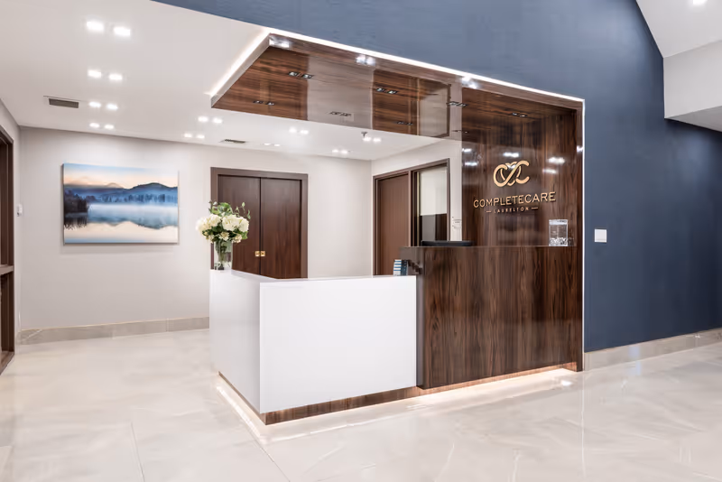 Reception area of Complete Care at Laurelton featuring a modern white and wood-paneled front desk with a vase of white flowers, a wall-mounted logo, a large landscape painting, and polished tile flooring.