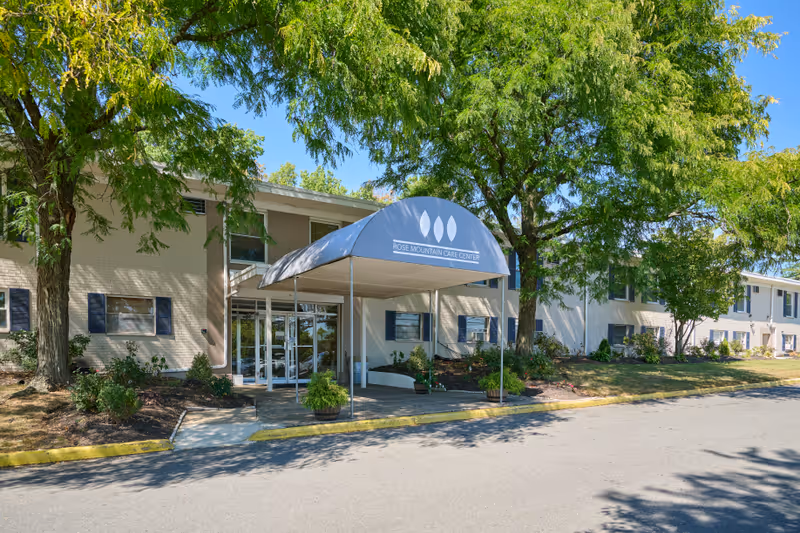 Exterior view of Rose Mountain Care Center showing the entrance with a blue awning displaying the facility's name. The building is two stories with several windows, surrounded by trees and landscaping under a clear blue sky.