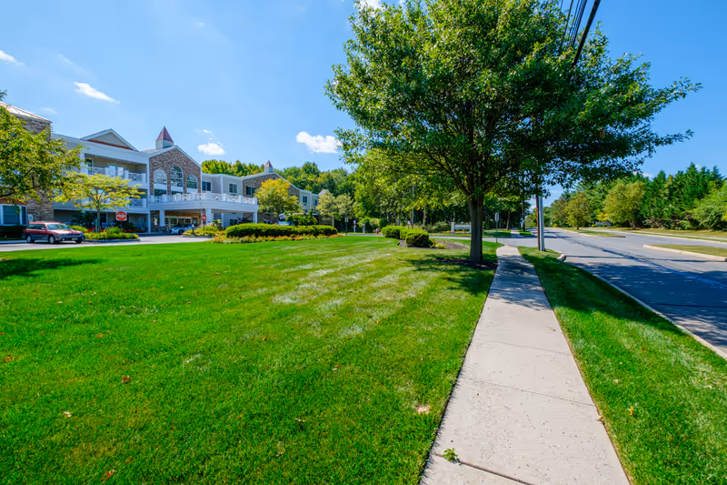 Exterior view of a senior living building with a large green lawn, sidewalk, and trees under a blue sky.