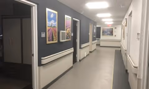 Interior corridor of a senior living facility with handrails, framed artwork, and open doorways under overhead lights.