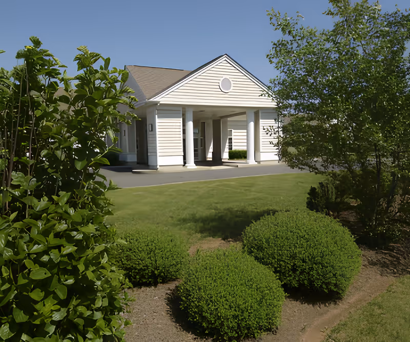 Exterior view of a white building with a covered entrance supported by four columns, surrounded by green bushes and trees under a clear blue sky.