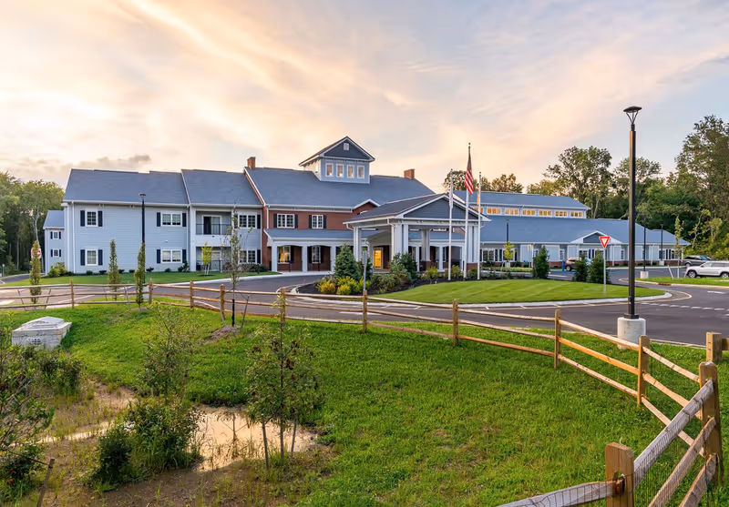 Exterior view of a large senior living facility building with a circular driveway, manicured lawn, wooden fence, and flagpoles with flags in front. The sky is partly cloudy with a warm sunset glow.
