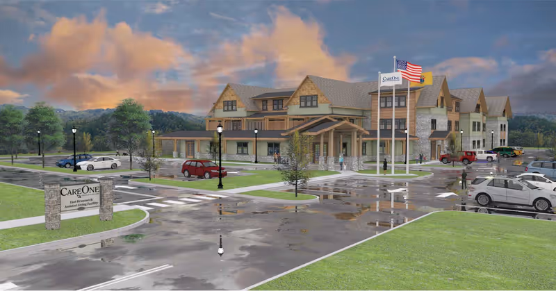 Exterior view of CareOne at East Brunswick Assisted Living facility with a large building, parking lot with several cars, flagpoles with American and other flags, and a sign at the entrance. The sky is partly cloudy with a sunset or sunrise glow.