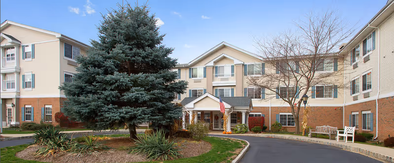 Exterior view of a senior living facility named Brookdale Wayne, showing a three-story building with beige siding and brick accents. There is a large evergreen tree and some landscaping in front of the entrance, which has a covered porch with an American flag. Benches and leafless trees are visible near the entrance.