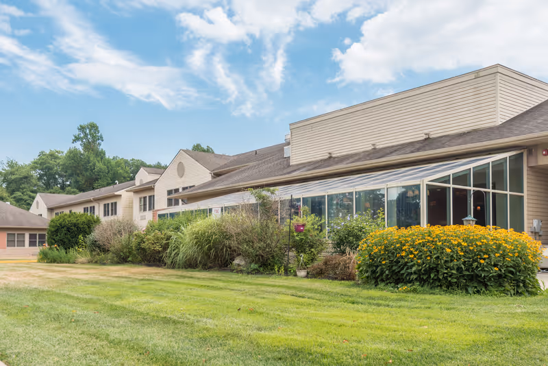 Exterior view of a senior living facility building with beige siding, large windows, and a well-maintained lawn with bushes and yellow flowers under a partly cloudy blue sky.