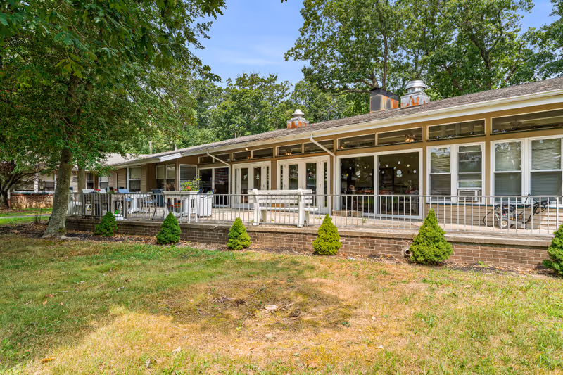 Exterior view of a single-story building with large windows and a patio area featuring outdoor seating and small shrubs in front. The building is surrounded by trees and grass under a clear sky.