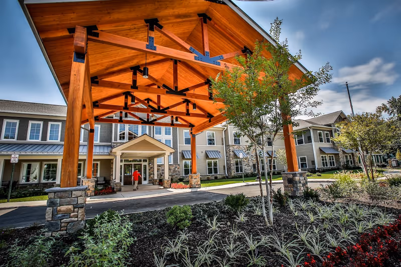 Entrance of a senior living facility with a large wooden canopy supported by stone pillars. The building has multiple windows and a person in a red shirt is walking towards the entrance. There are landscaped plants and trees in the foreground under a partly cloudy sky.