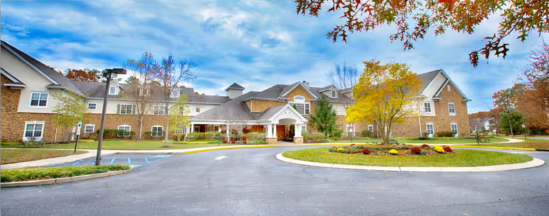 Front exterior view of a large senior living facility building with a circular driveway, landscaped garden with colorful flowers, and trees with autumn foliage under a partly cloudy sky.