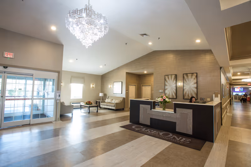 Bright and spacious reception area of a senior living facility with a modern chandelier, a front desk with a receptionist, a seating area with a sofa and chairs, and large glass entrance doors.