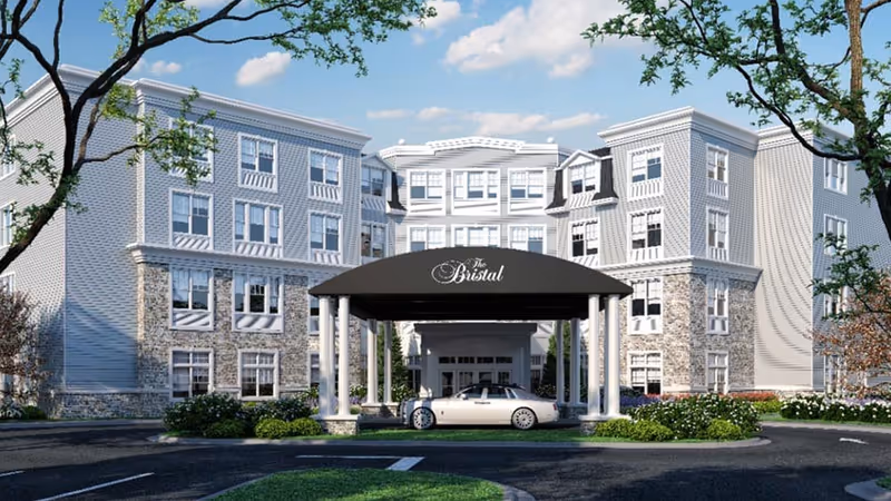 Front exterior view of The Bristal Assisted Living at Wayne building with a covered entrance, a white car parked under the canopy, surrounded by trees and landscaping under a blue sky with some clouds.