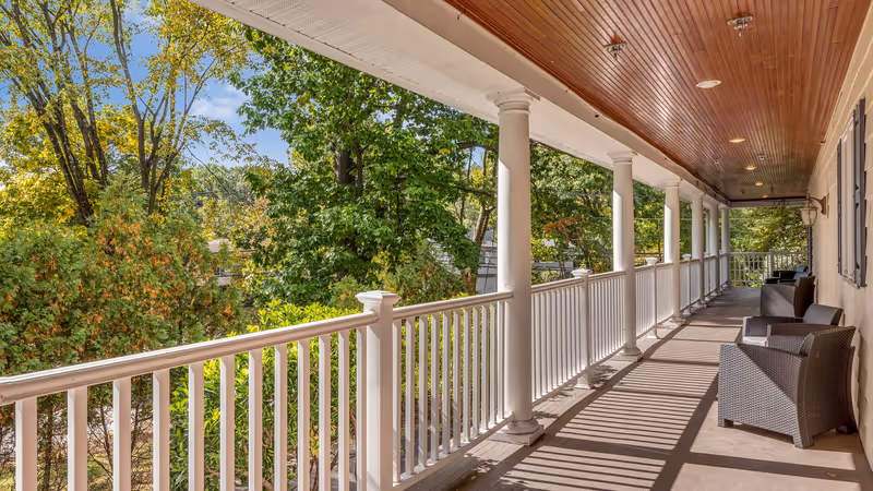 Covered porch with white columns, railing and wicker chairs overlooking trees.