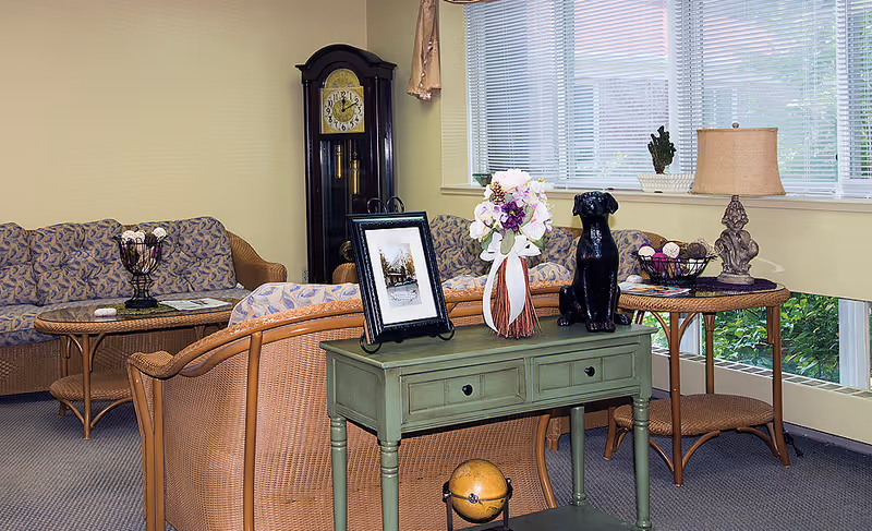 A cozy living room area with wicker furniture including a sofa and chairs with patterned cushions. A green console table holds a framed photo, a floral arrangement, and a black dog statue. There is a tall grandfather clock in the corner and a side table with a lamp and decorative items near a window with blinds.