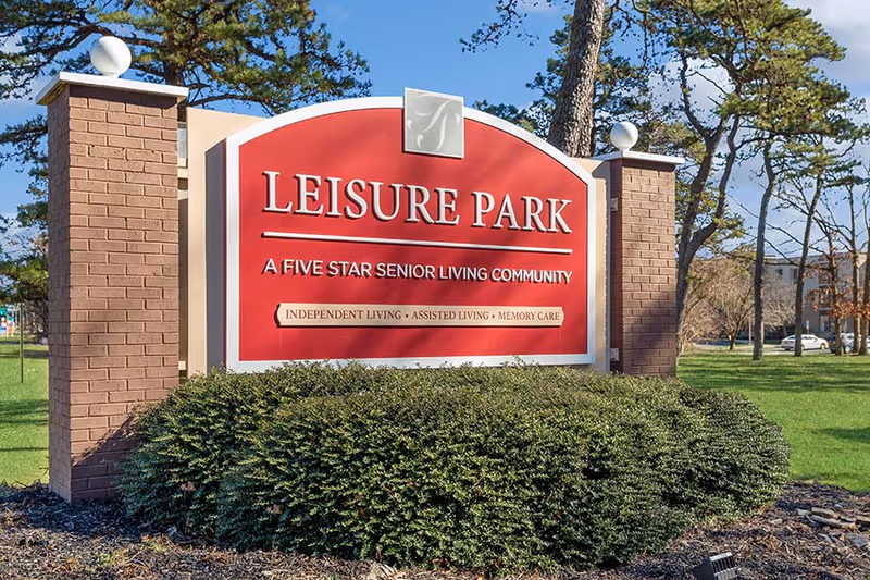 A red entrance sign reading "Leisure Park" and "A Five Star Senior Living Community" mounted between brick pillars with shrubs and trees behind it.