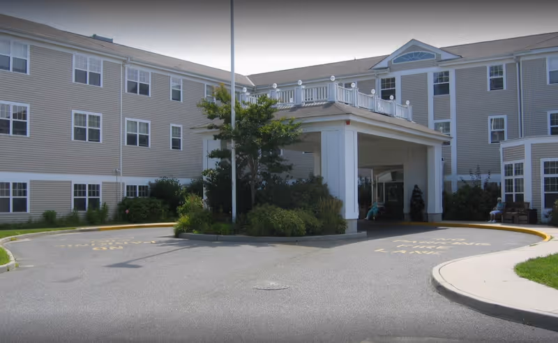 Exterior view of a multi-story assisted living facility with beige siding and white trim. The building has many windows and a covered entrance with a circular driveway. There are some bushes and trees near the entrance, and a person sitting on a bench to the right.