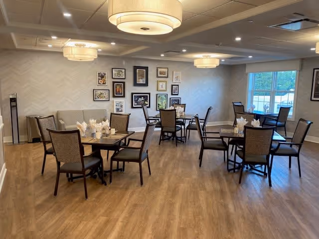 A dining room in a senior living facility with several square tables, each surrounded by four chairs. The tables are set with napkins and cups. The room has wooden flooring, beige walls with a subtle pattern, and a gallery wall of framed pictures. Large windows allow natural light to enter, and there are modern ceiling light fixtures.